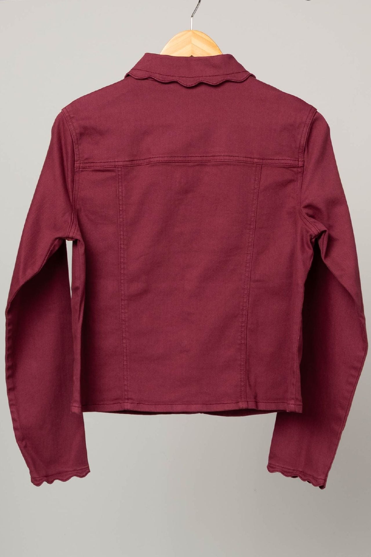 A maroon long-sleeve cropped shirt jacket with scalloped edges on the collar and cuffs, hanging on a wooden hanger against a plain light gray background.
