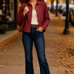 A woman stands on an autumn street wearing a burgundy button-up shirt jacket over a cream top, dark blue flare jeans, and brown ankle boots, with a brown shoulder bag.