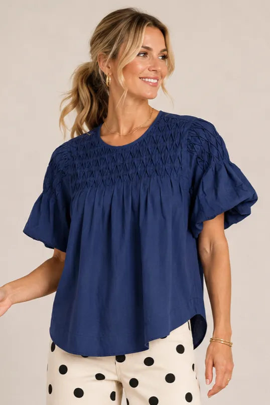 A woman wearing a navy blue short-sleeve blouse with a smocked bodice and ruffled sleeves, paired with cream pants featuring black polka dots, against a plain beige studio background.