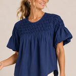 A woman wearing a navy blue short-sleeve blouse with a smocked bodice and ruffled sleeves, paired with cream pants featuring black polka dots, against a plain beige studio background.