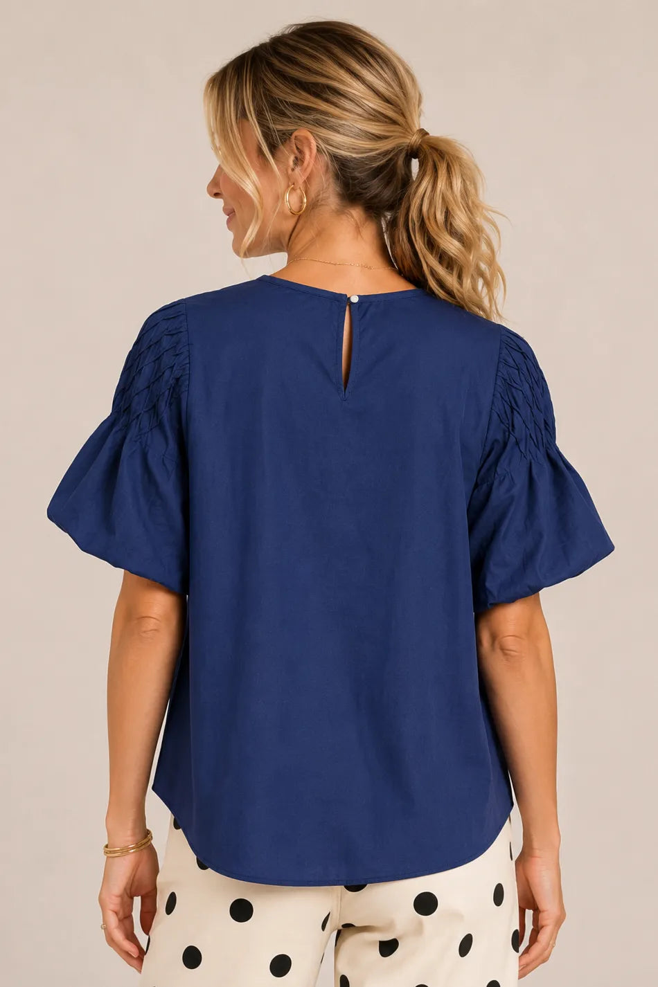 A woman shown from the back wearing a navy blue short-sleeve blouse with ruffled sleeves and a small keyhole cutout at the back, paired with cream pants with black polka dots, against a plain beige studio background.