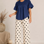 A woman modeling a navy blue smocked short-sleeve blouse and white wide-leg pants with black polka dots, standing in a styled indoor setting with a neutral wall, woven rug, and decorative vase.