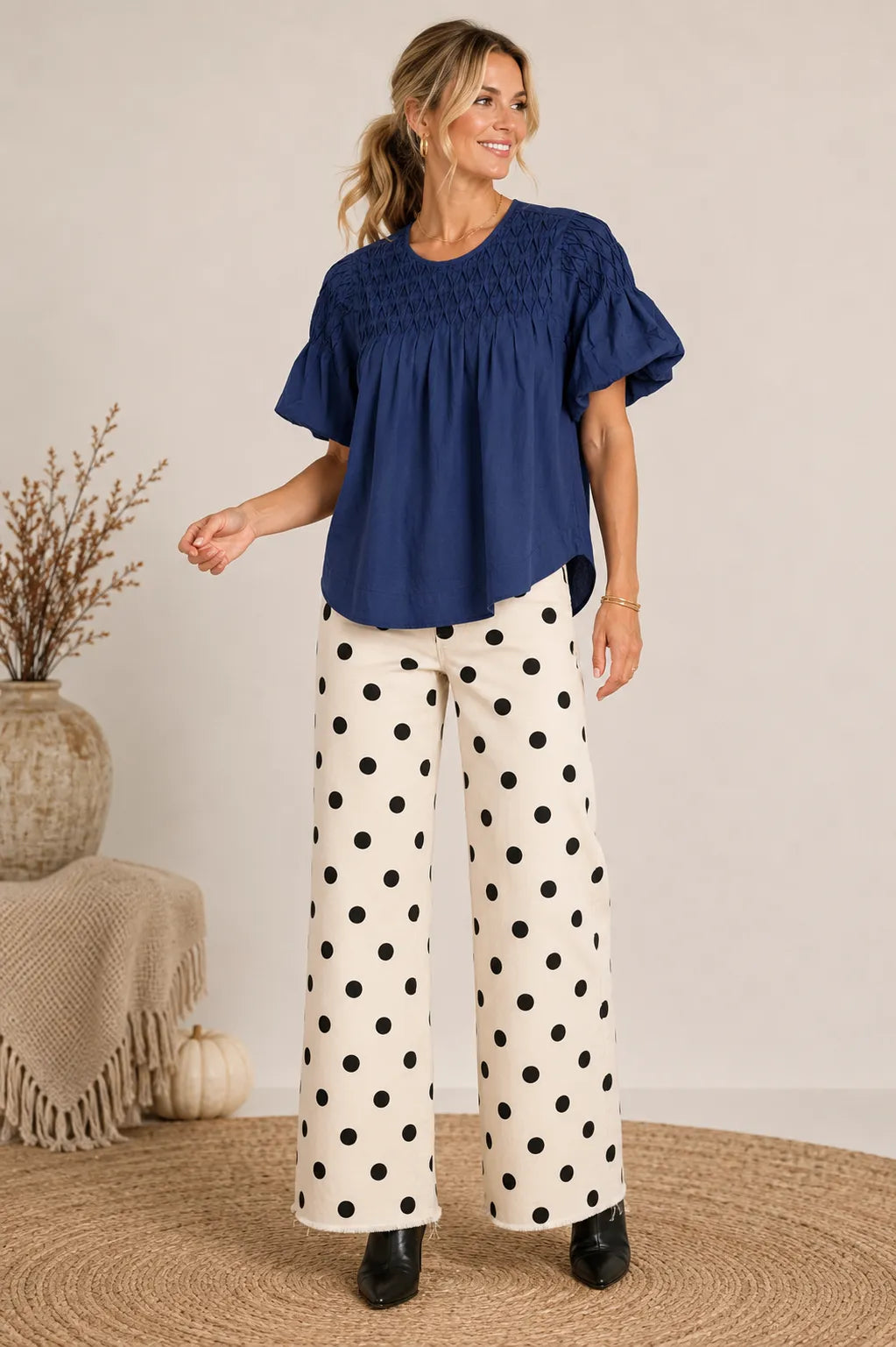 A woman modeling a navy blue smocked short-sleeve blouse and white wide-leg pants with black polka dots, standing in a styled indoor setting with a neutral wall, woven rug, and decorative vase.