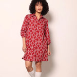 A model with curly black hair stands against a plain off-white wall wearing a red collared shirt dress with a white geometric pattern, paired with white knee-high socks and white sneakers.