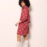 A model with curly black hair is shown from the side wearing a red and white patterned shirt dress, white socks, and white platform sneakers against a plain off-white wall.