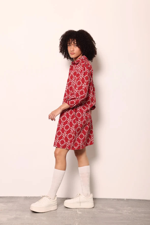 A model with curly black hair is shown from the side wearing a red and white patterned shirt dress, white socks, and white platform sneakers against a plain off-white wall.