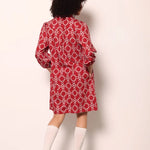 A child model shown from the back wearing a red and white geometric patterned long-sleeve shirt dress, paired with white knee-high socks and white sneakers, standing against a plain off-white wall.
