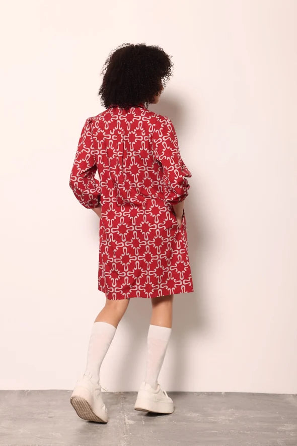 A child model shown from the back wearing a red and white geometric patterned long-sleeve shirt dress, paired with white knee-high socks and white sneakers, standing against a plain off-white wall.