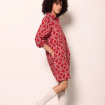 A model with curly black hair wearing a red and white patterned shirt dress, white socks, and white sneakers, standing against a plain off-white wall.