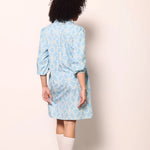 A person shown from the back wearing a light blue patterned shirt dress with three-quarter sleeves, paired with white socks and white sneakers, standing against a plain off-white wall.