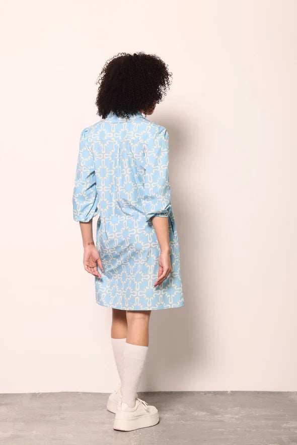 A person shown from the back wearing a light blue patterned shirt dress with three-quarter sleeves, paired with white socks and white sneakers, standing against a plain off-white wall.