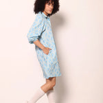 A model with curly black hair is shown from the side wearing a light blue and white patterned shirt dress, white knee-high socks, and white sneakers, standing against a plain off-white wall.