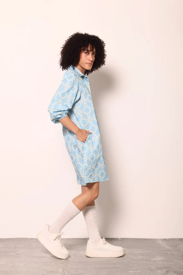 A model with curly black hair is shown from the side wearing a light blue and white patterned shirt dress, white knee-high socks, and white sneakers, standing against a plain off-white wall.