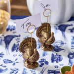 Two small metallic gold place settings shaped like turkeys holding white name cards on a blue-and-white patterned tablecloth.