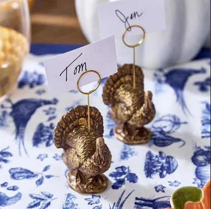 Two small metallic gold place settings shaped like turkeys holding white name cards on a blue-and-white patterned tablecloth.