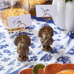Two small metallic turkey-shaped place card holders on a blue-and-white patterned tablecloth, each holding a white name card with the names Tom and Jen.