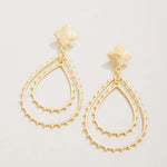 A pair of gold-tone dangle earrings with a small flower-shaped stud and two layered teardrop-shaped wire frames outlined with tiny bead details, shown on a light off-white background.