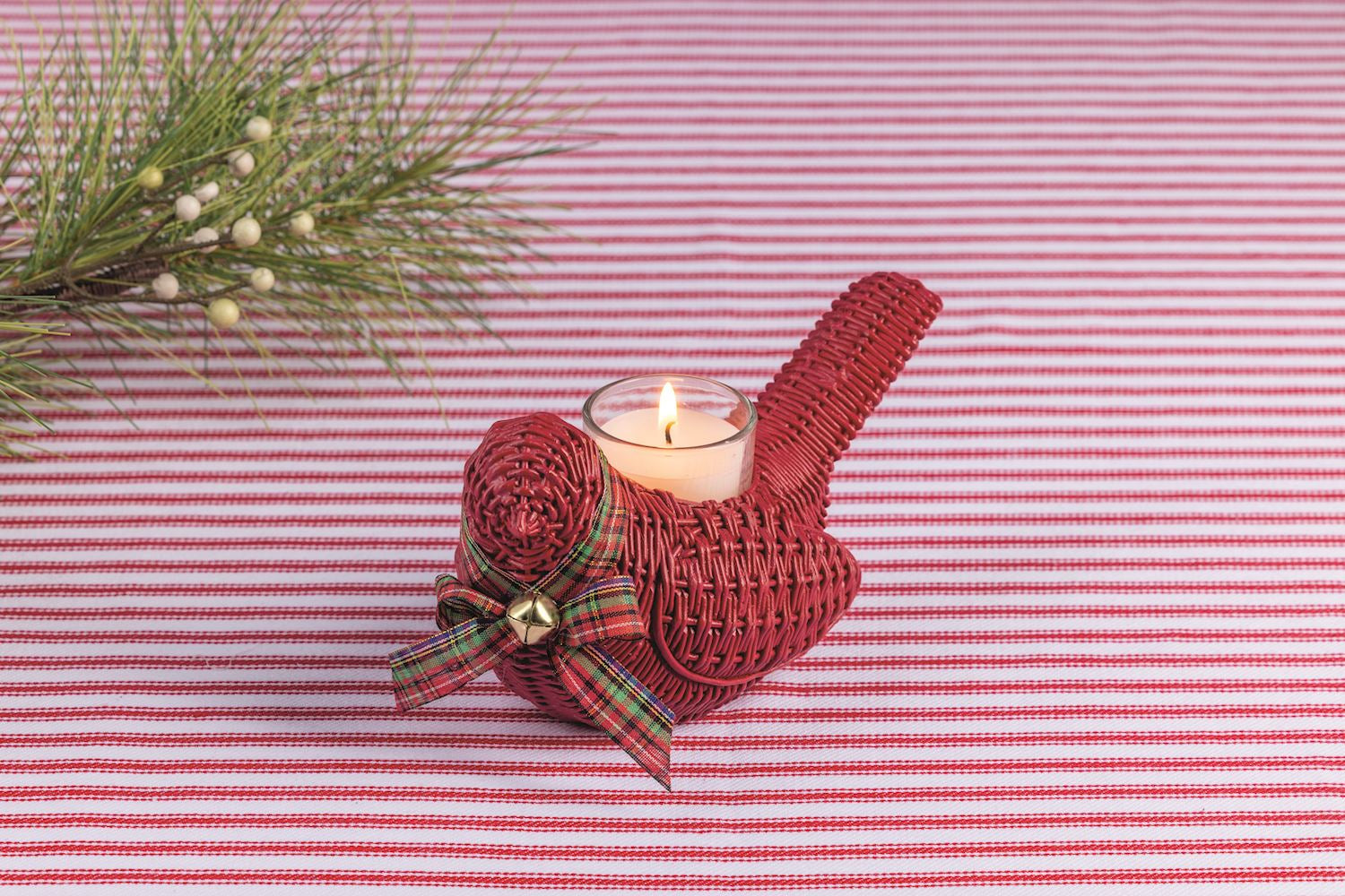 A red woven bird-shaped candle holder with a lit white candle inside, decorated with a red plaid ribbon and small gold bell, set on a red and white striped fabric background with pine branches in the corner.