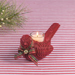 A red woven bird-shaped candle holder with a lit white candle inside, decorated with a red plaid ribbon and small gold bell, set on a red and white striped fabric background with pine branches in the corner.