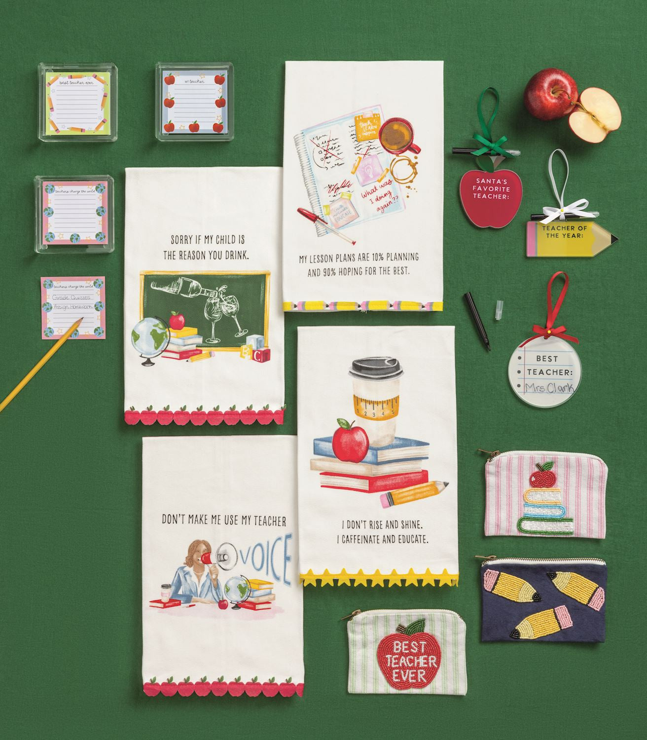 A flat-lay display of various school-themed paper goods and small accessories on a dark green background. The collection includes greeting cards, small note cards, a coffee mug tag, a 'Best Teacher Ever' ornament, a 'Don't Make Me Que My Teacher' card, a 'Don't Make Me Que My Teacher' pouch, and other matching school-themed items.