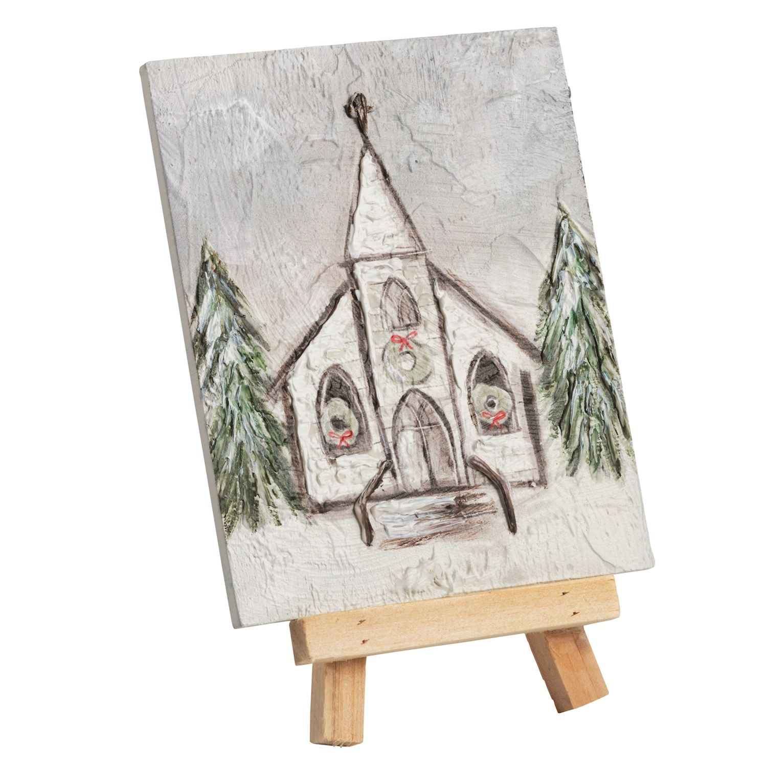 Hand-painted wooden Christmas church canvas print displayed on a small wooden easel. The textured off-white canvas features a hand-drawn church with green pine trees and a red wreath, set against a plain white background.