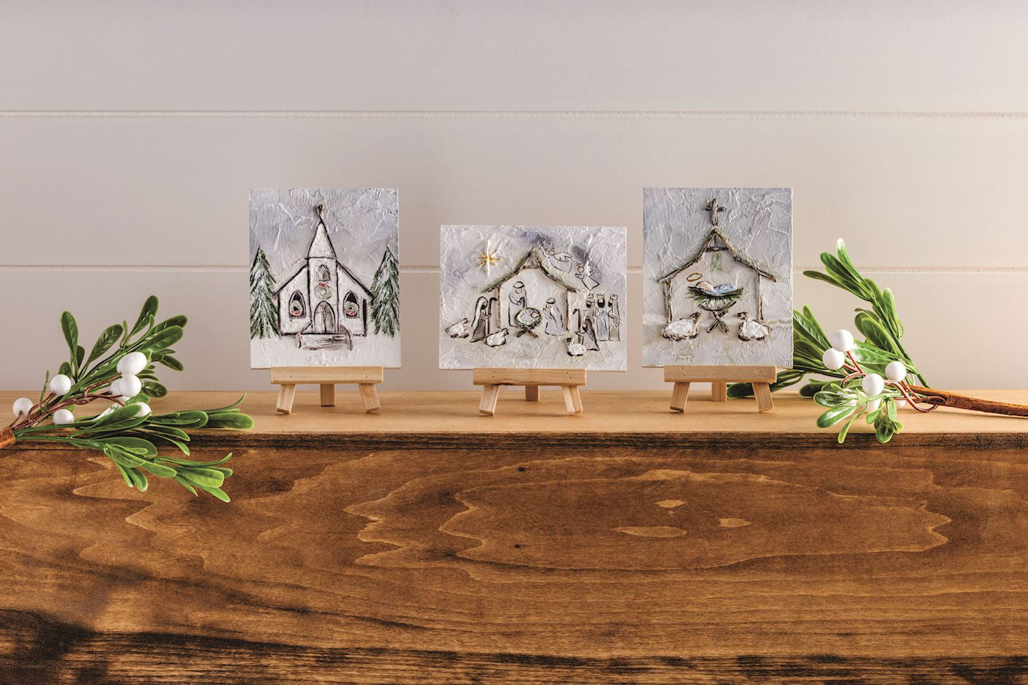 Three small square wall art prints on mini easels, each showing a different winter holiday scene: a church, a nativity stable, and a stable with animals. The prints are displayed on a wooden surface with greenery and white berries against a light paneled wall.