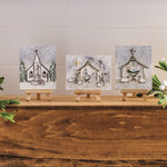 Three small square wall art prints on mini easels, each showing a different winter holiday scene: a church, a nativity stable, and a stable with animals. The prints are displayed on a wooden surface with greenery and white berries against a light paneled wall.