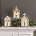 Three decorative church-shaped ornaments with gold and white detailing, displayed on a white mantel with green garland against a dark gray wall.