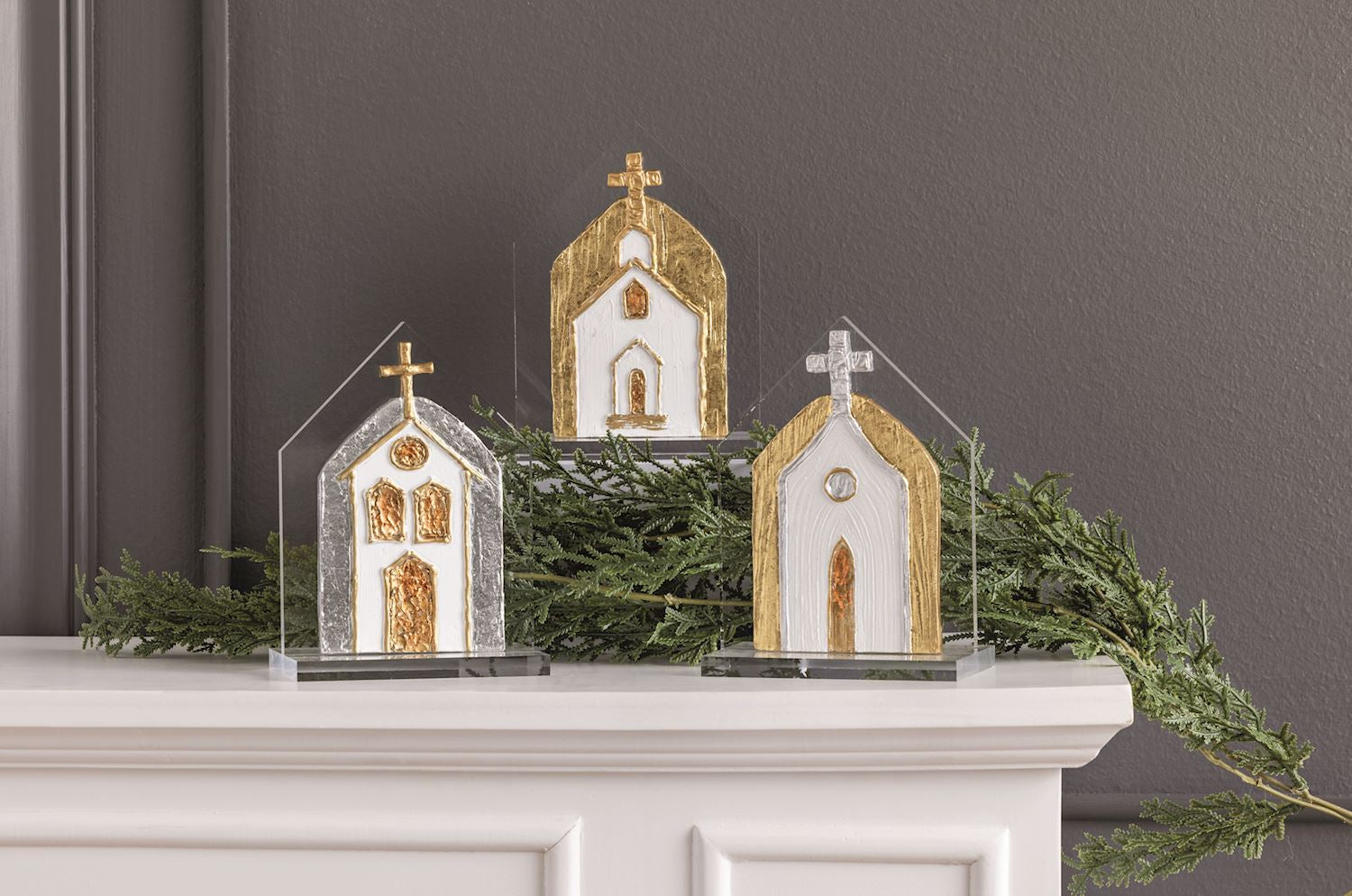 Three decorative church-shaped ornaments with gold and white detailing, displayed on a white mantel with green garland against a dark gray wall.