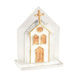 A small decorative church-shaped object with a gold cross on top, featuring textured gold and white detailing, displayed on a clear acrylic block against a white background.