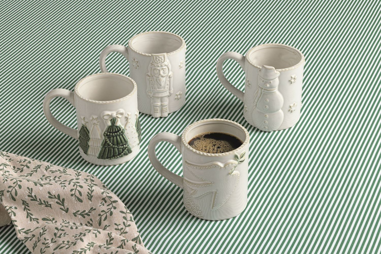 Four white ceramic mugs with embossed holiday illustrations, arranged on a green and white striped fabric background. One mug in the foreground contains coffee, and a matching patterned cloth is partially visible in the lower left corner.
