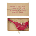 A red bird-shaped ornament with white floral embroidery and a small black cardinal head, displayed in an open kraft gift box with a hanging string. The box lid above reads, 'Forever in your heart, a loved one so dear.