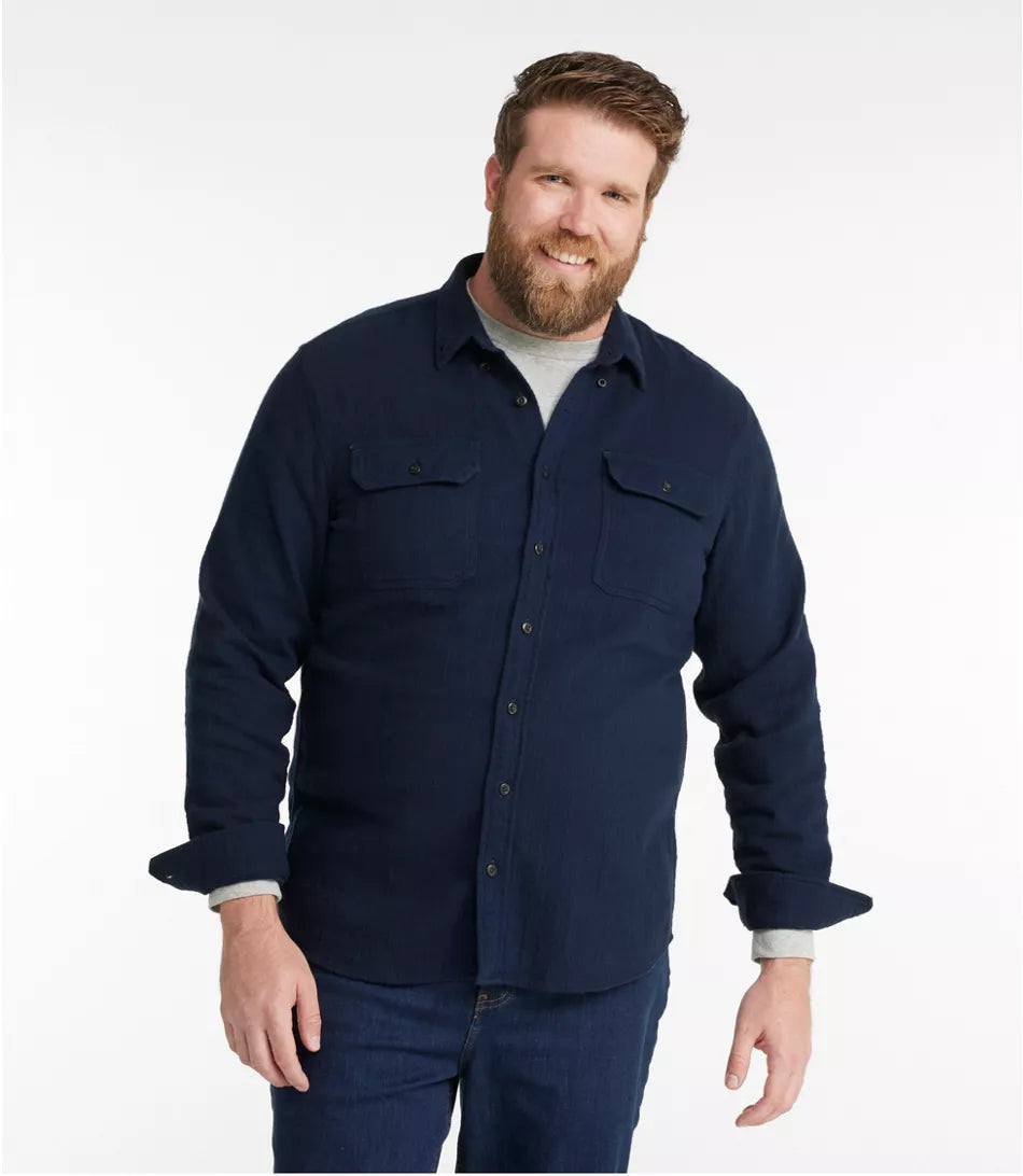 A man wearing a dark navy blue long-sleeve button-up shirt with two chest pockets, layered over a light gray t-shirt and paired with dark blue jeans, against a plain white studio background.