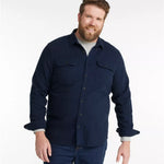 A man wearing a dark navy blue long-sleeve button-up shirt with two chest pockets, layered over a light gray t-shirt and paired with dark blue jeans, against a plain white studio background.