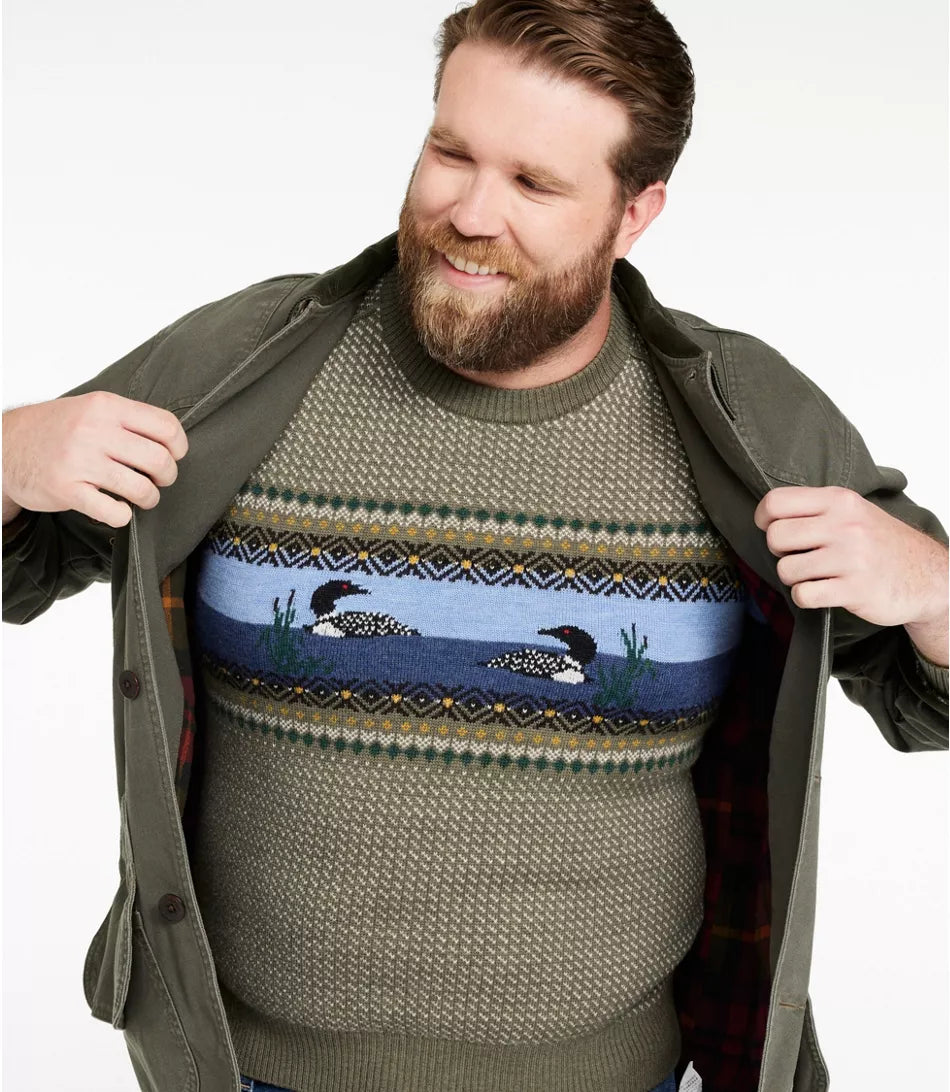 A smiling bearded man wearing a knit crewneck sweater with a colorful horizontal pattern of loons and birds, opening an olive green jacket to show the sweater underneath, against a plain white background.