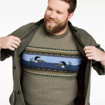 A smiling bearded man wearing a knit crewneck sweater with a colorful horizontal pattern of loons and birds, opening an olive green jacket to show the sweater underneath, against a plain white background.