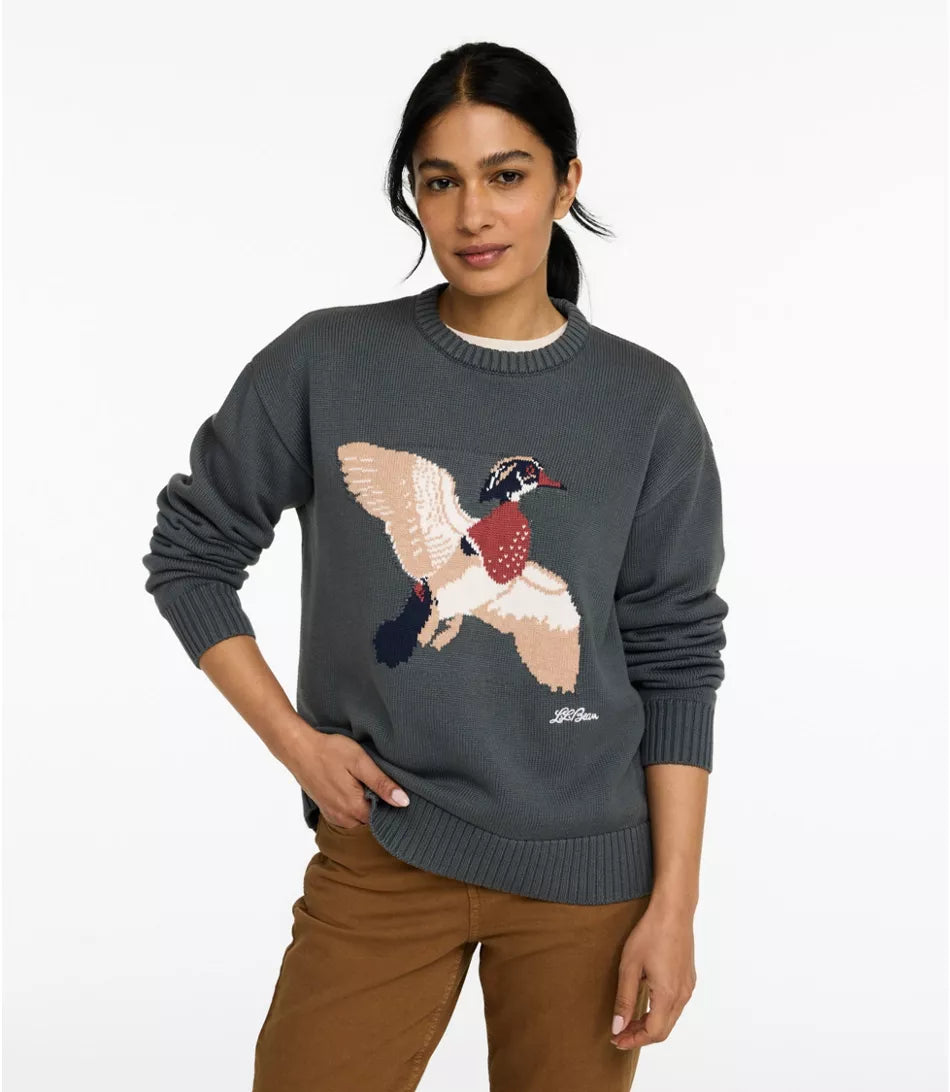 A woman wearing a dark gray knit crewneck sweater with a large embroidered pheasant graphic on the front, paired with brown pants, against a plain white background.