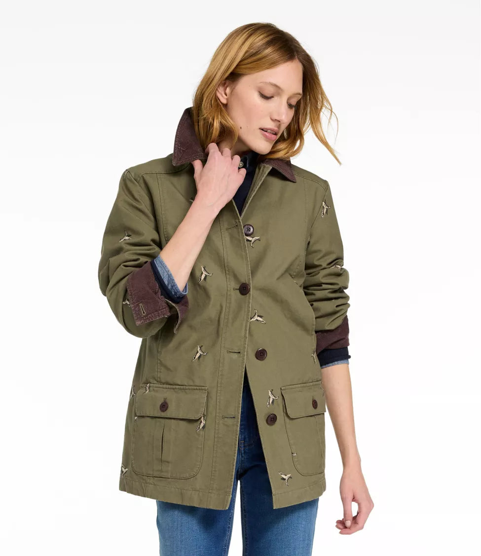 A woman wearing an olive green utility jacket with dark brown corduroy collar and cuffs, featuring small embroidered details, paired with blue jeans against a white studio background.