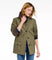 A woman wearing an olive green utility jacket with dark brown corduroy collar and cuffs, featuring small embroidered details, paired with blue jeans against a white studio background.