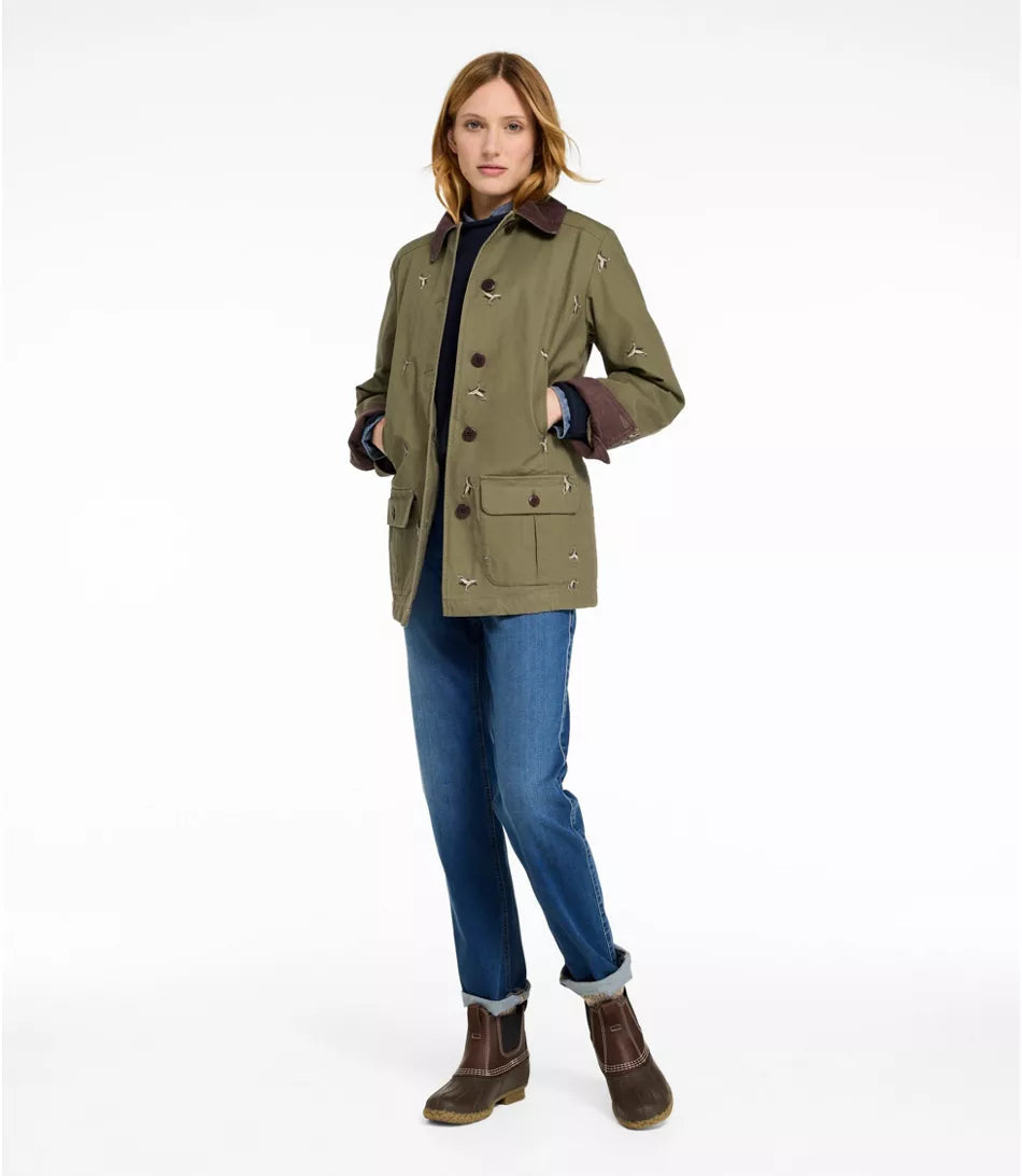 A woman modeling an olive green waxed canvas jacket with brown leather-like trim and buttons, paired with blue jeans and brown boots, against a plain white studio background.