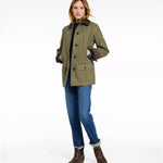 A woman modeling an olive green waxed canvas jacket with brown leather-like trim and buttons, paired with blue jeans and brown boots, against a plain white studio background.