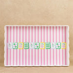 A rectangular serving tray with scalloped edges, featuring a pink and white vertical stripe pattern and a row of illustrated mahjong tiles across the center, set against a textured beige background.