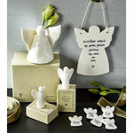 A set of white ceramic angel figurines and hanging plaques with printed remembrance messages, displayed with their boxes on a dark tabletop against a gray wall. One larger angel figure stands on a box with green buds in its hand, while smaller angel figures are arranged in front, including one on a small base and one hanging plaque that reads, 'Guardian angels up above please protect the ones we love'.