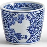 A white ceramic cup or small planter decorated with blue floral and botanical patterns in an oval medallion design, shown on a light gray background.