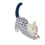 A ceramic figurine of a stretching cat in a white glaze, decorated with a blue floral pattern, featuring a dark blue tail and small gold eyes and nose.