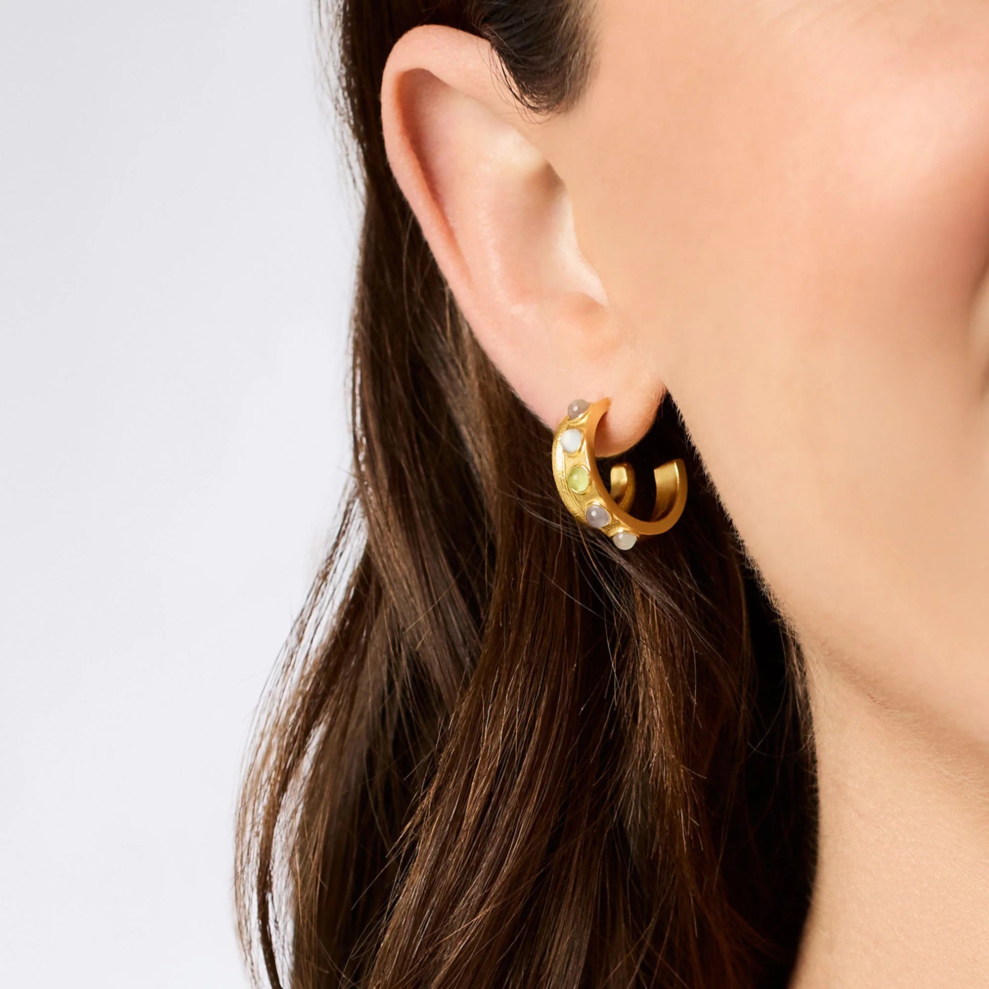 A close-up of a model's ear wearing a gold crescent-shaped hoop earring adorned with small clear stones, set against a light gray background.