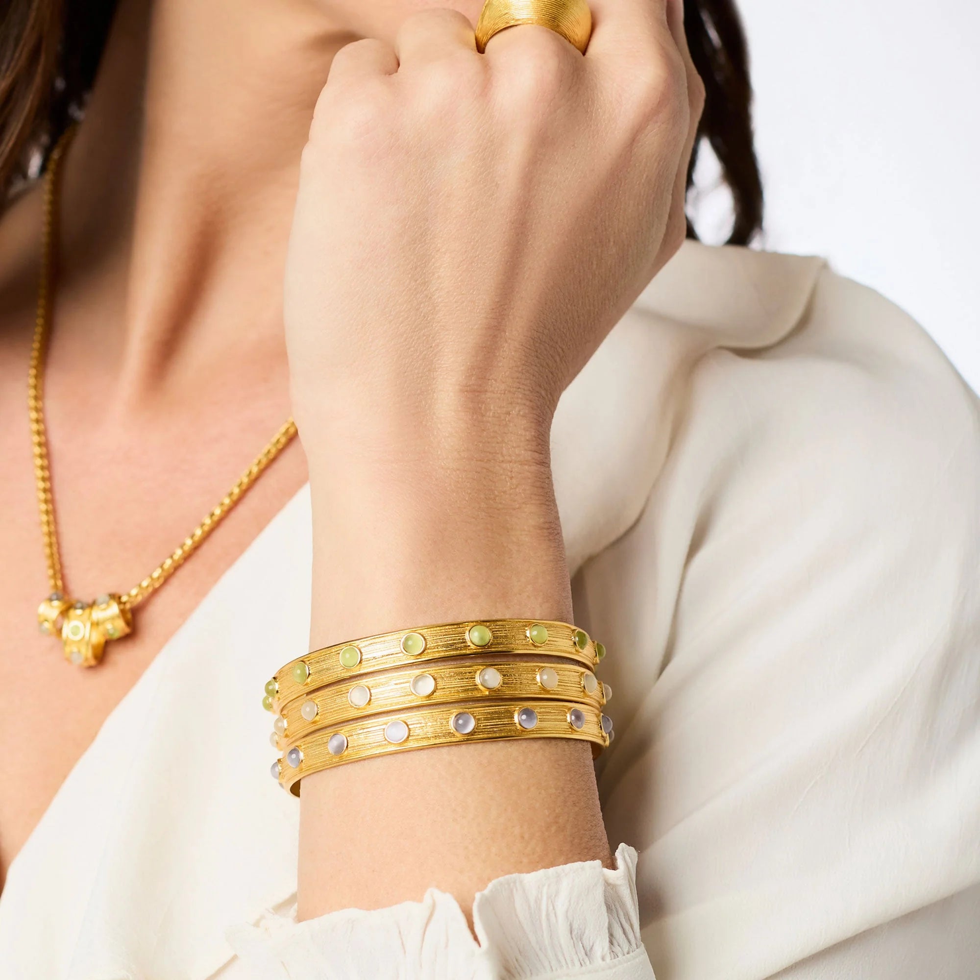A close-up of a person wearing a stack of three gold bangle bracelets on their wrist, each decorated with small round green and white stones. The person is also wearing a gold necklace and a gold ring, and is dressed in a white blouse.