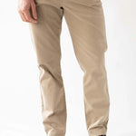 A man wearing beige slim-fit chino pants, a blue t-shirt, and brown leather lace-up shoes, standing against a white studio background.