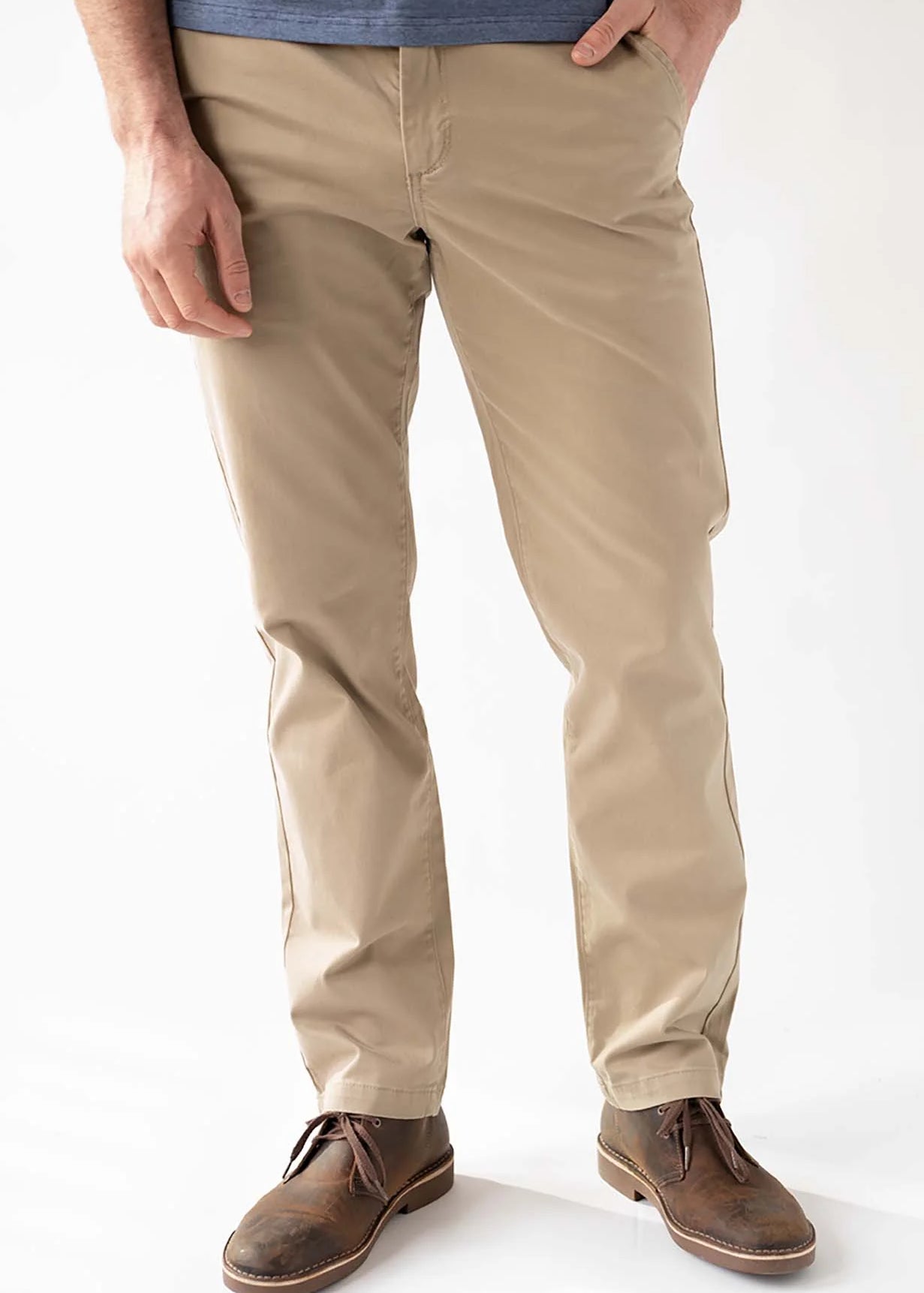 A man wearing beige slim-fit chino pants, a blue t-shirt, and brown leather lace-up shoes, standing against a white studio background.
