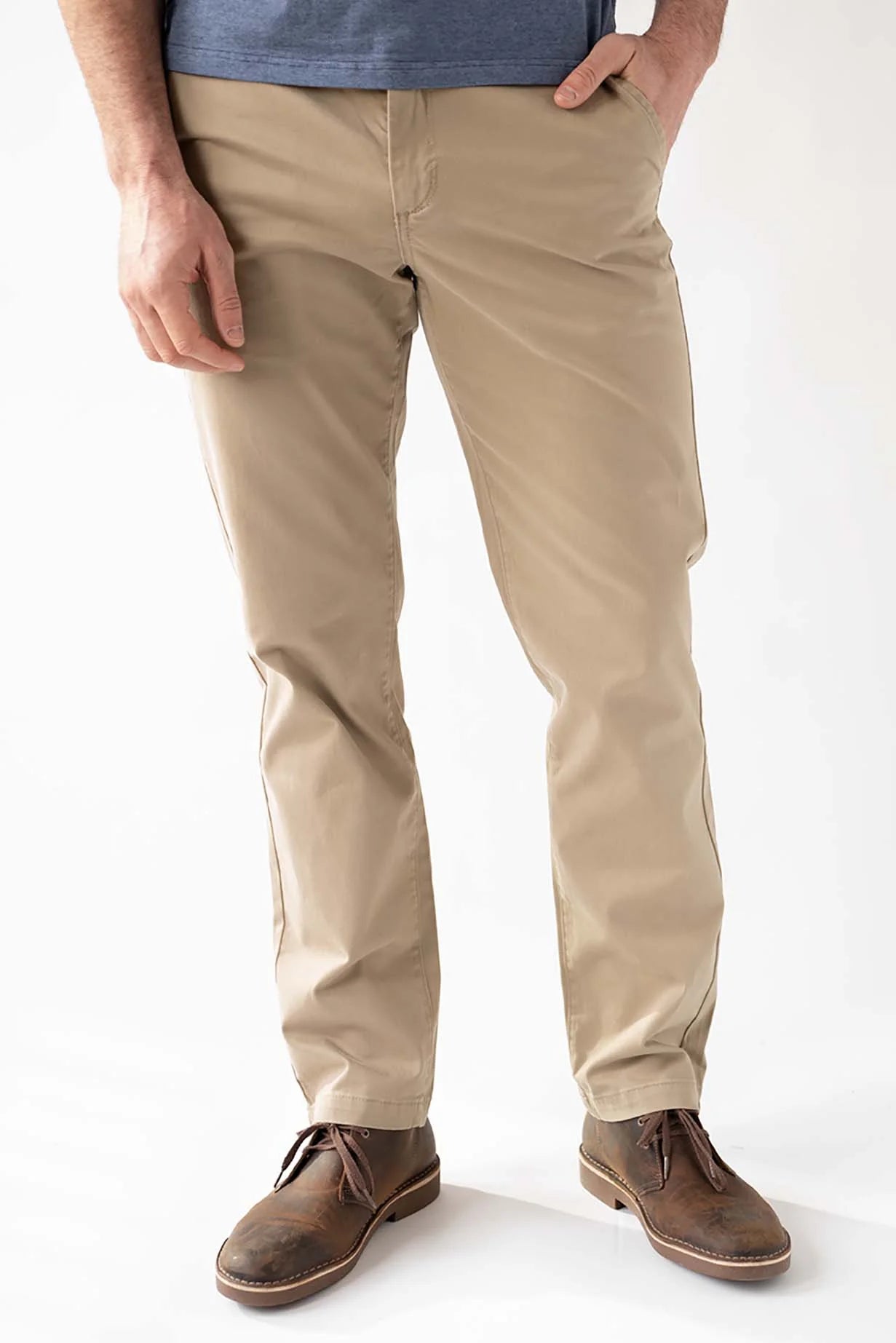 A man wearing beige slim-fit chino pants, a blue t-shirt, and brown leather lace-up shoes, standing against a white studio background.